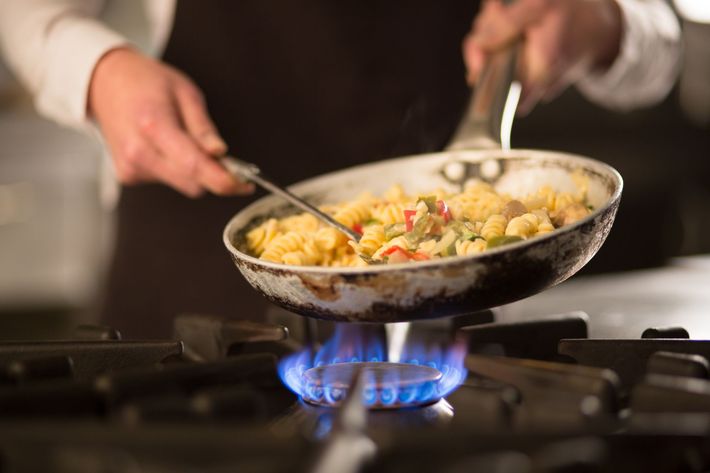 Chef stir-frying vegetables and pasta in a pan over a gas flame.