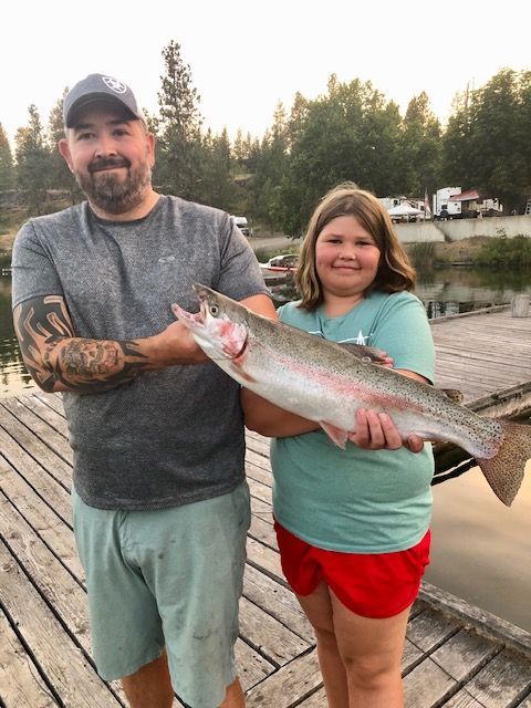 Man and child holding a large rainbow trout on a wooden dock near water, with a campground in the background.