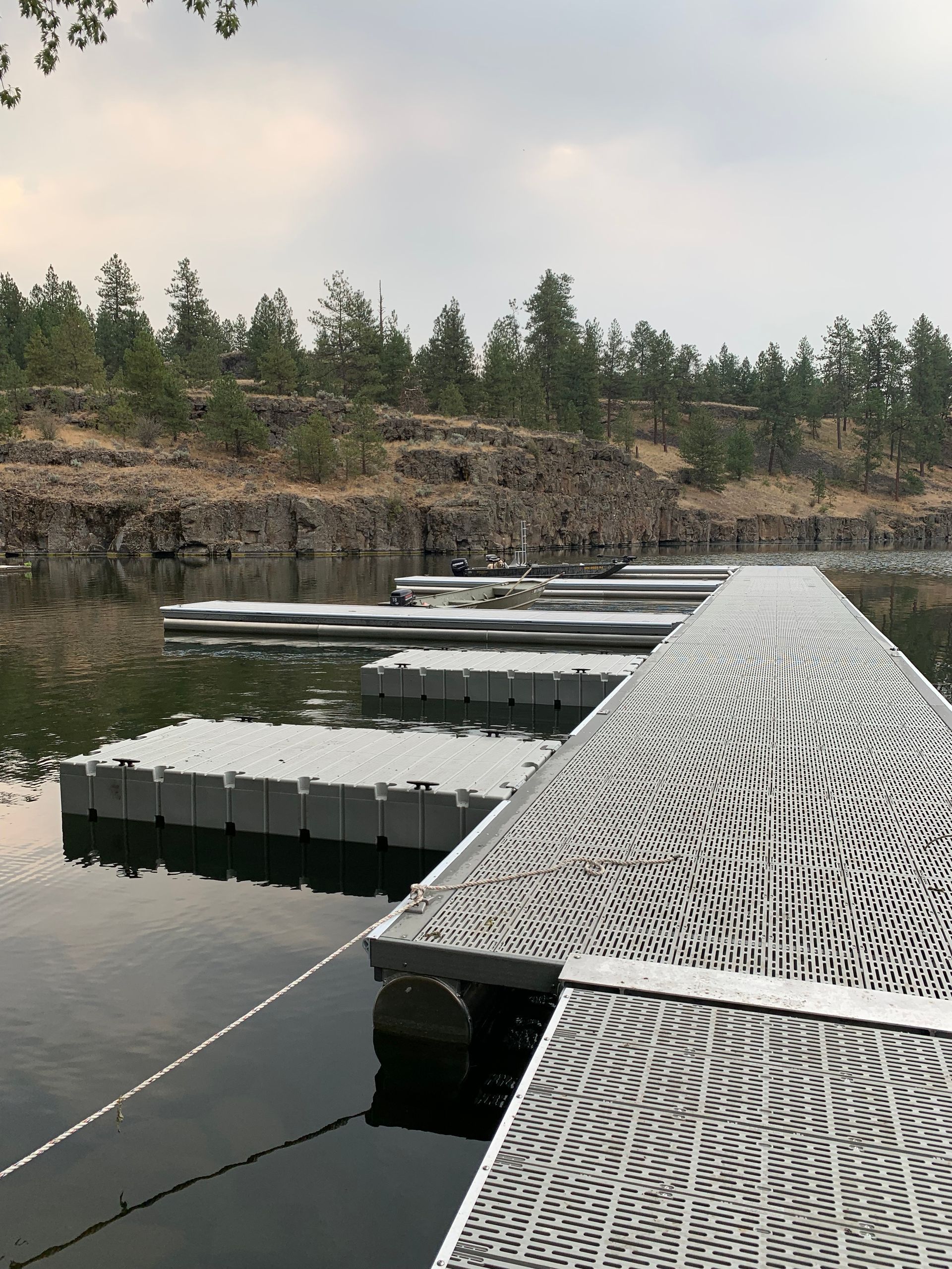 Floating dock with patterned surface on calm water; trees on rocky shoreline.