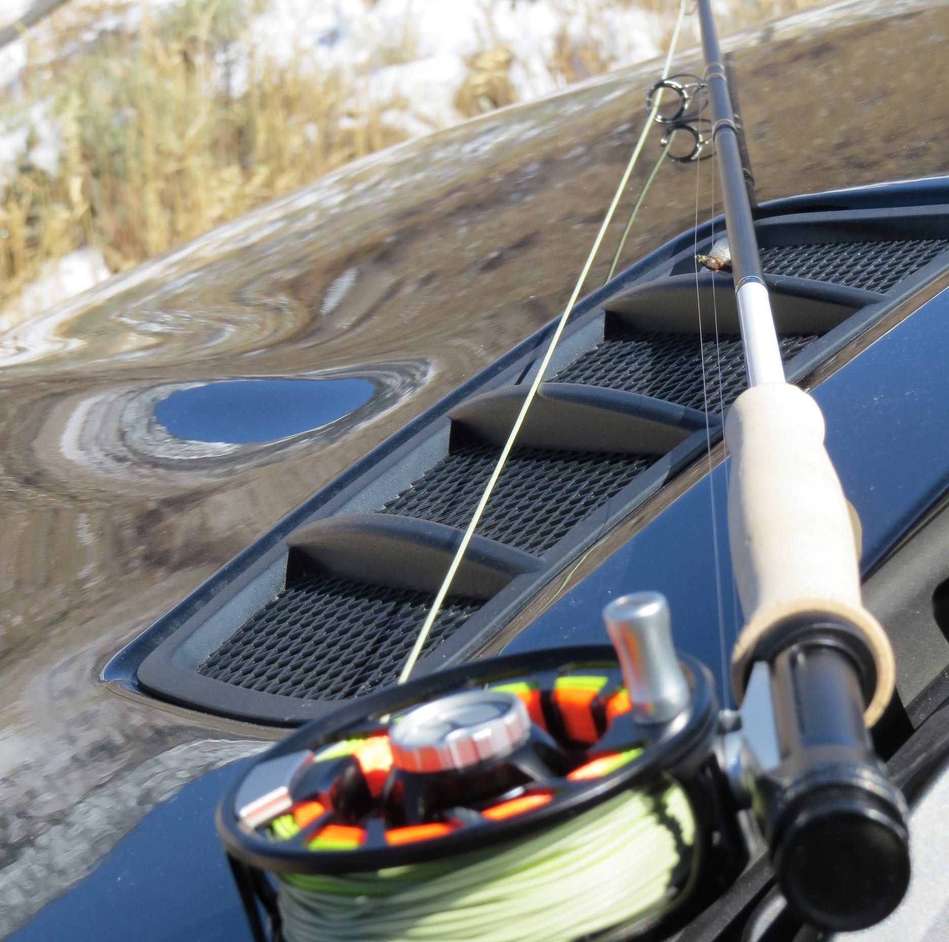 Fishing rod and reel resting on a black vehicle hood with a vent, likely preparing for fishing.