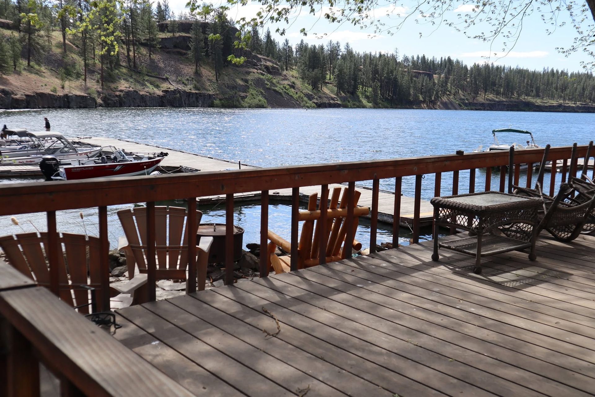 Deck overlooking a lake with Adirondack chairs, a small table, and a dock. Trees line the far shore.