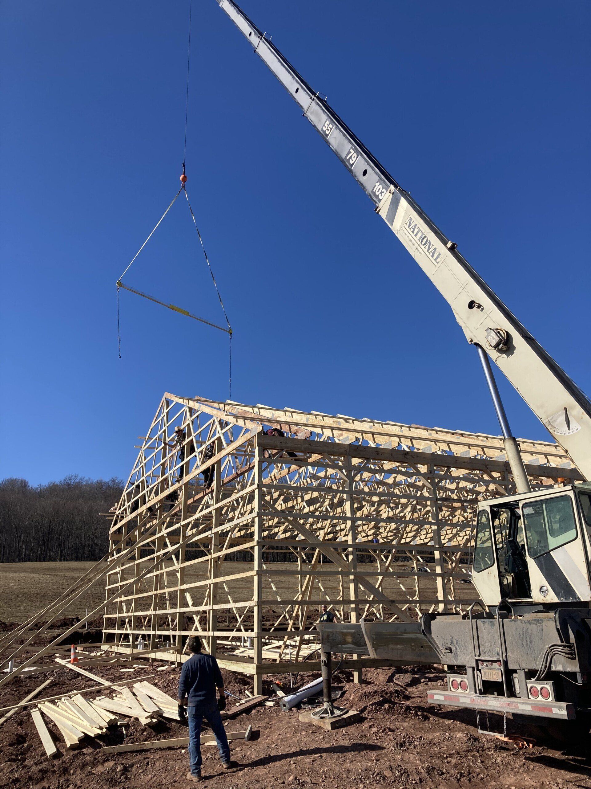A crane lifting a beam onto a partially built wooden barn frame on a sunny day. Man watches.