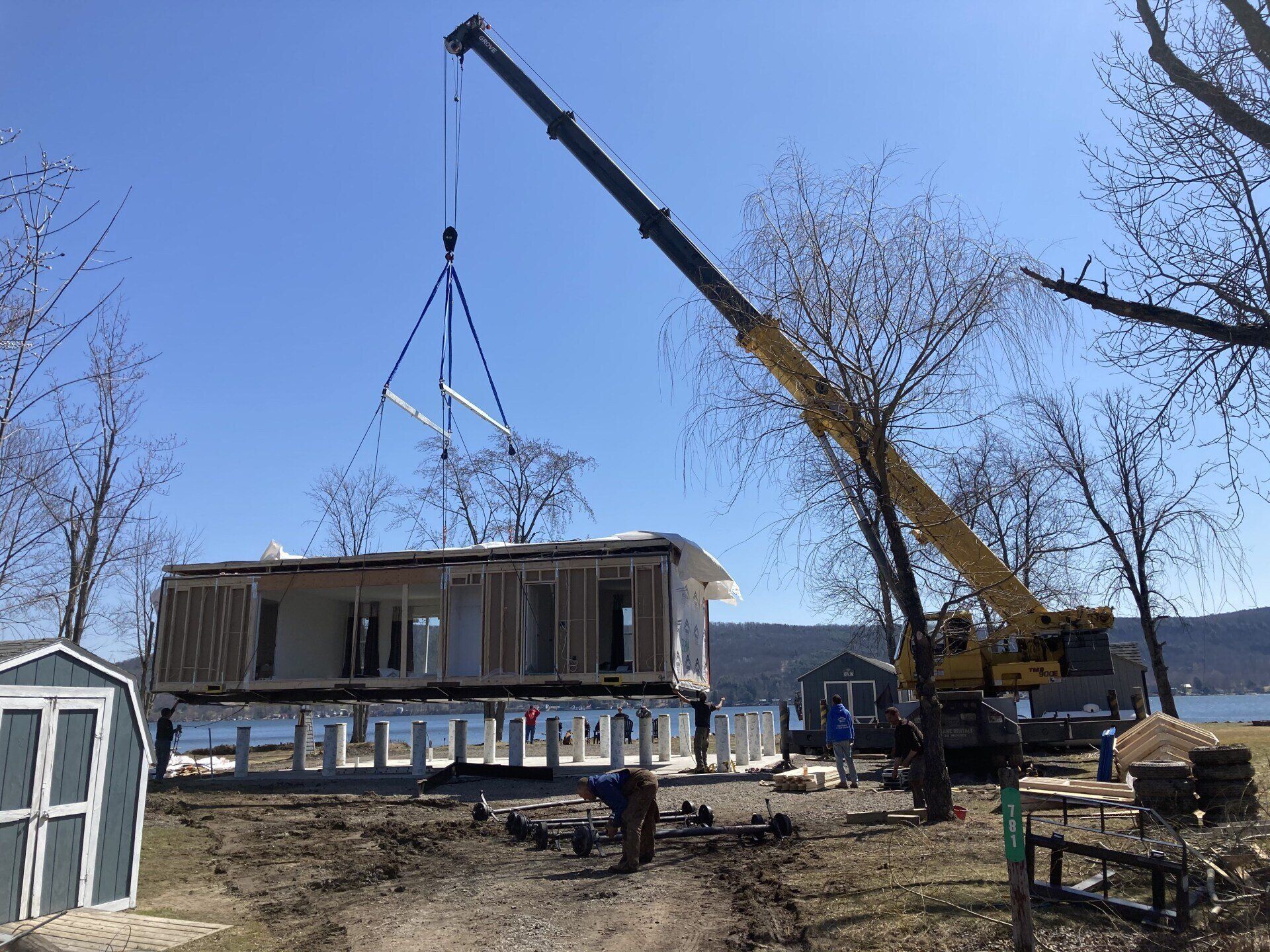A crane lifting a house frame over wooden piers by a body of water on a sunny day.