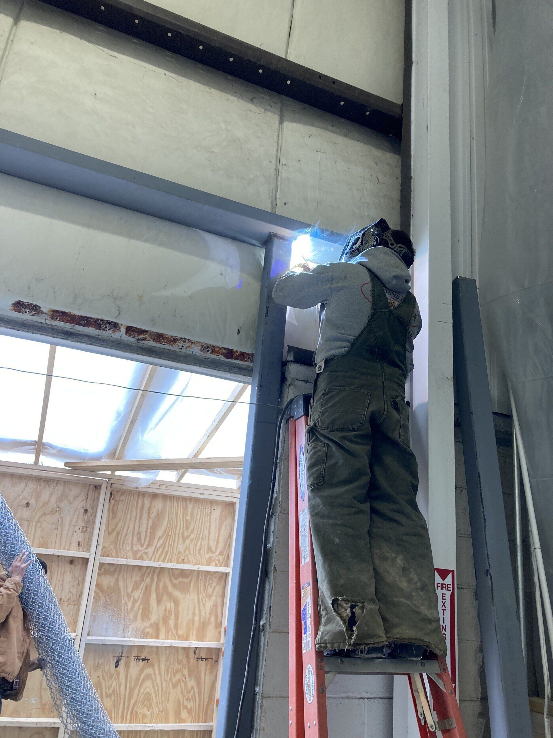 Welder in overalls on a ladder, welding metal frame in a building.