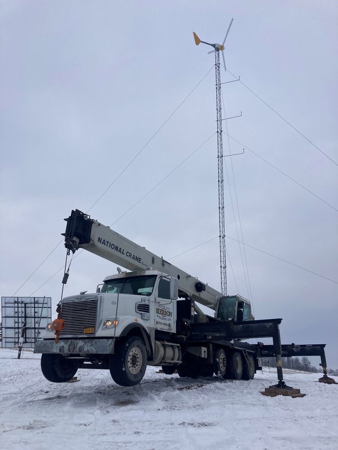 A crane truck installing a small wind turbine in a snowy field on a cloudy day.