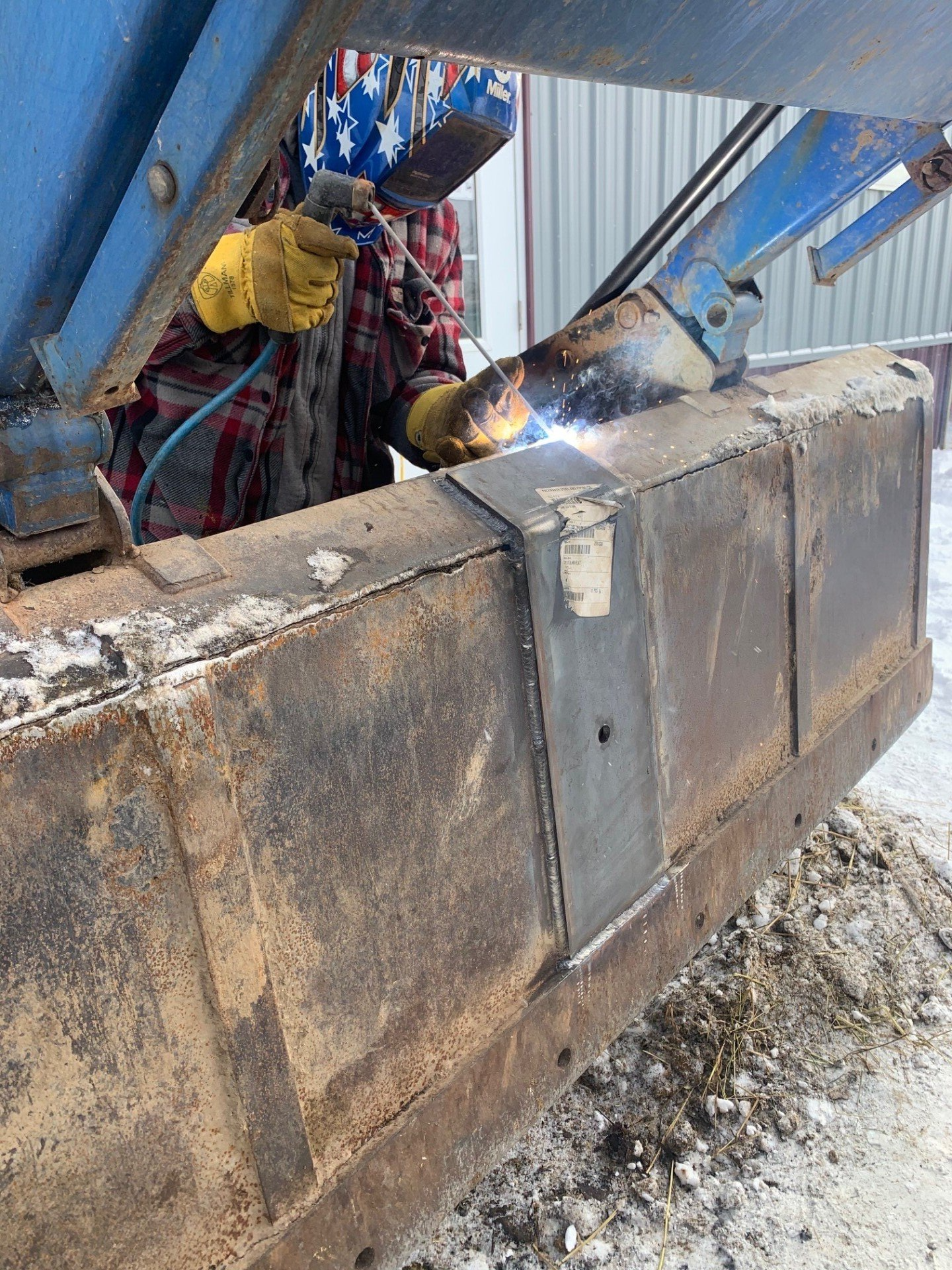 Welder with star-spangled helmet working on a rusty metal bucket in a snowy outdoor setting.