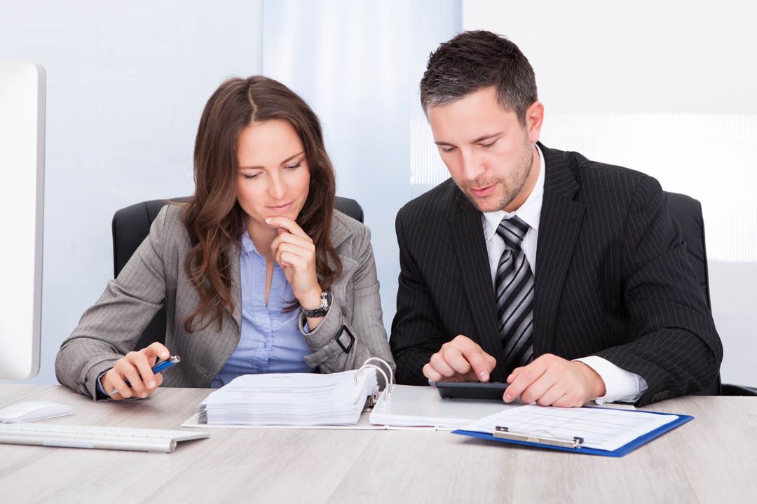 Two coworkers reviewing paperwork at a desk, one writing and the other reading a clipboard.
