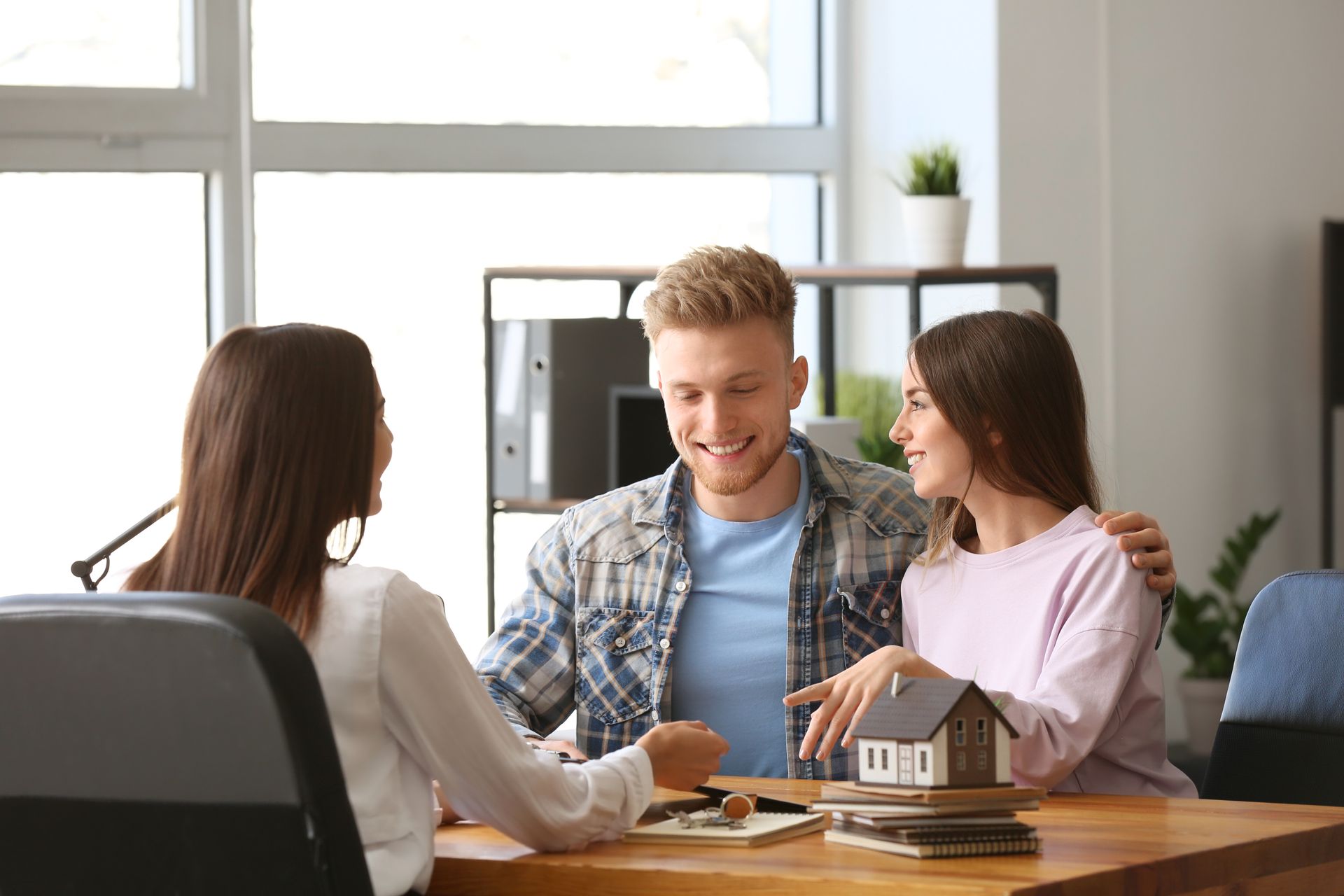 Three people meeting at a table with a house model, discussing a home purchase in a bright office.