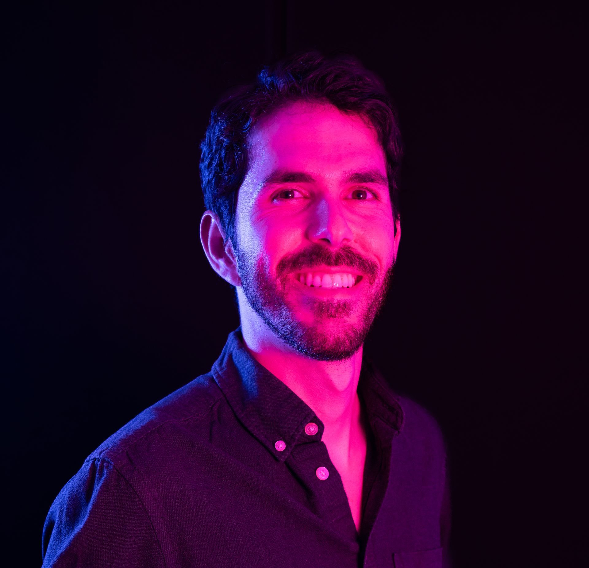 Portrait of a smiling person with dark hair, lit by vibrant pink and blue studio lights on a black background