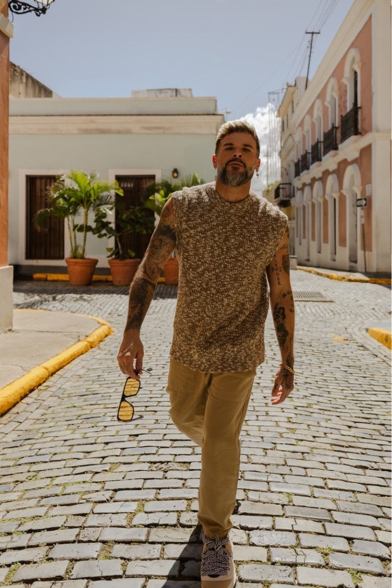 Man walking down a sunlit cobblestone street, holding sunglasses, with buildings and potted plants nearby.