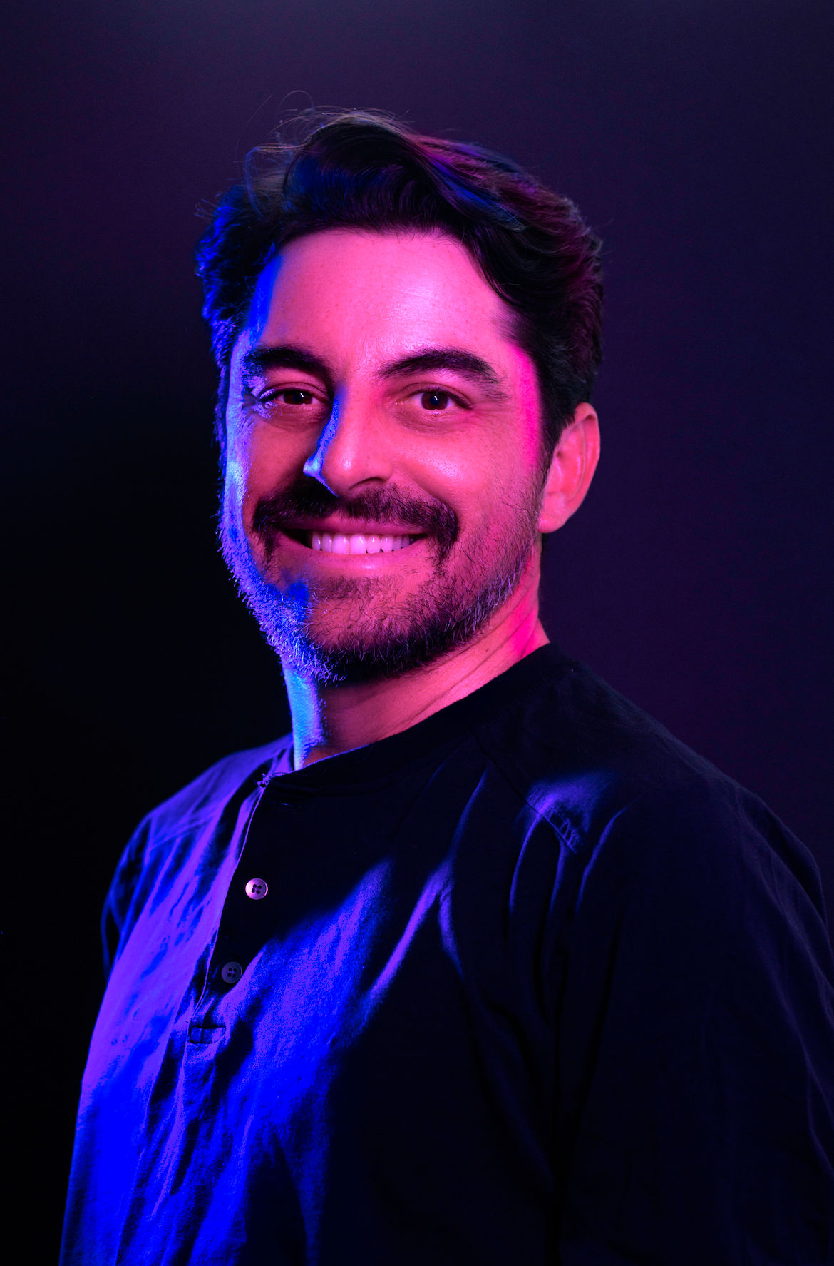 Portrait of a smiling person in a black shirt, lit with purple and blue studio lighting