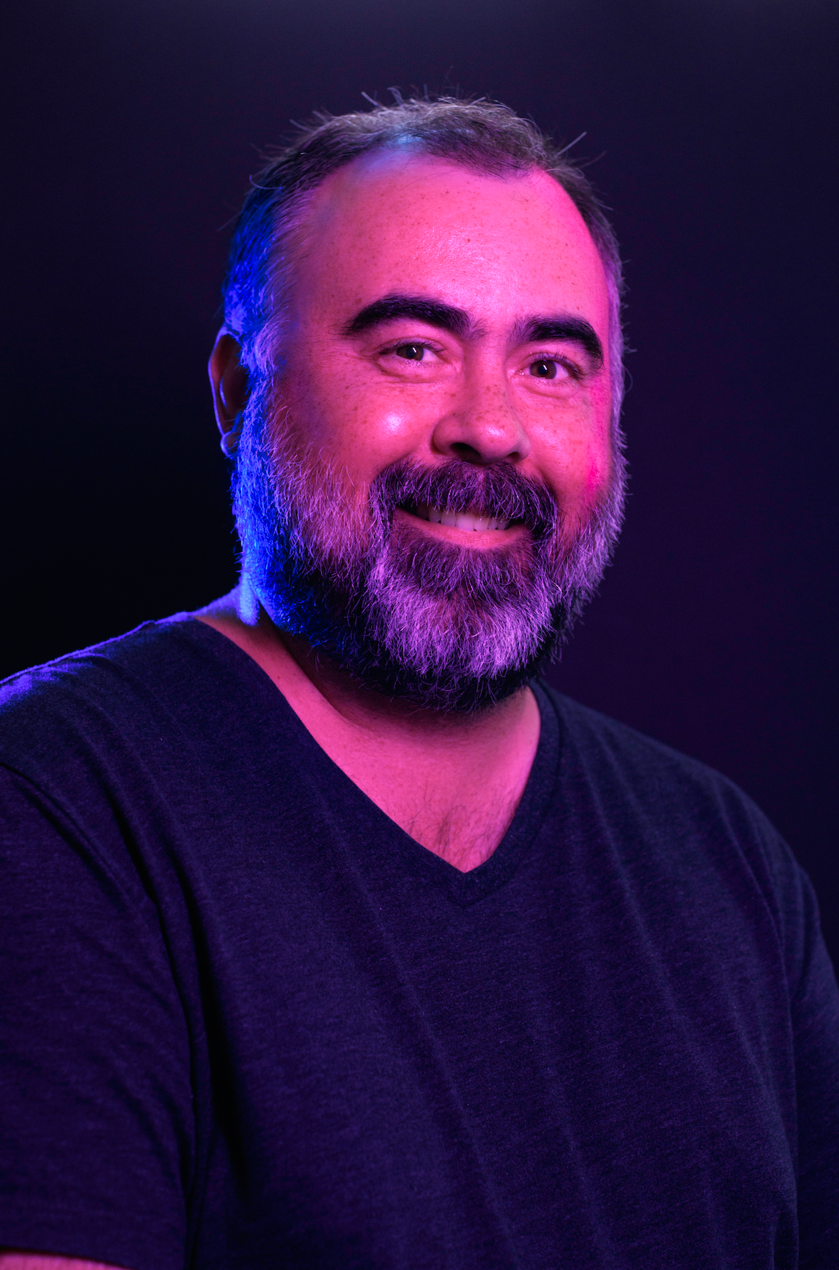 Portrait of a smiling bearded man in a dark shirt, lit by purple and blue studio lights