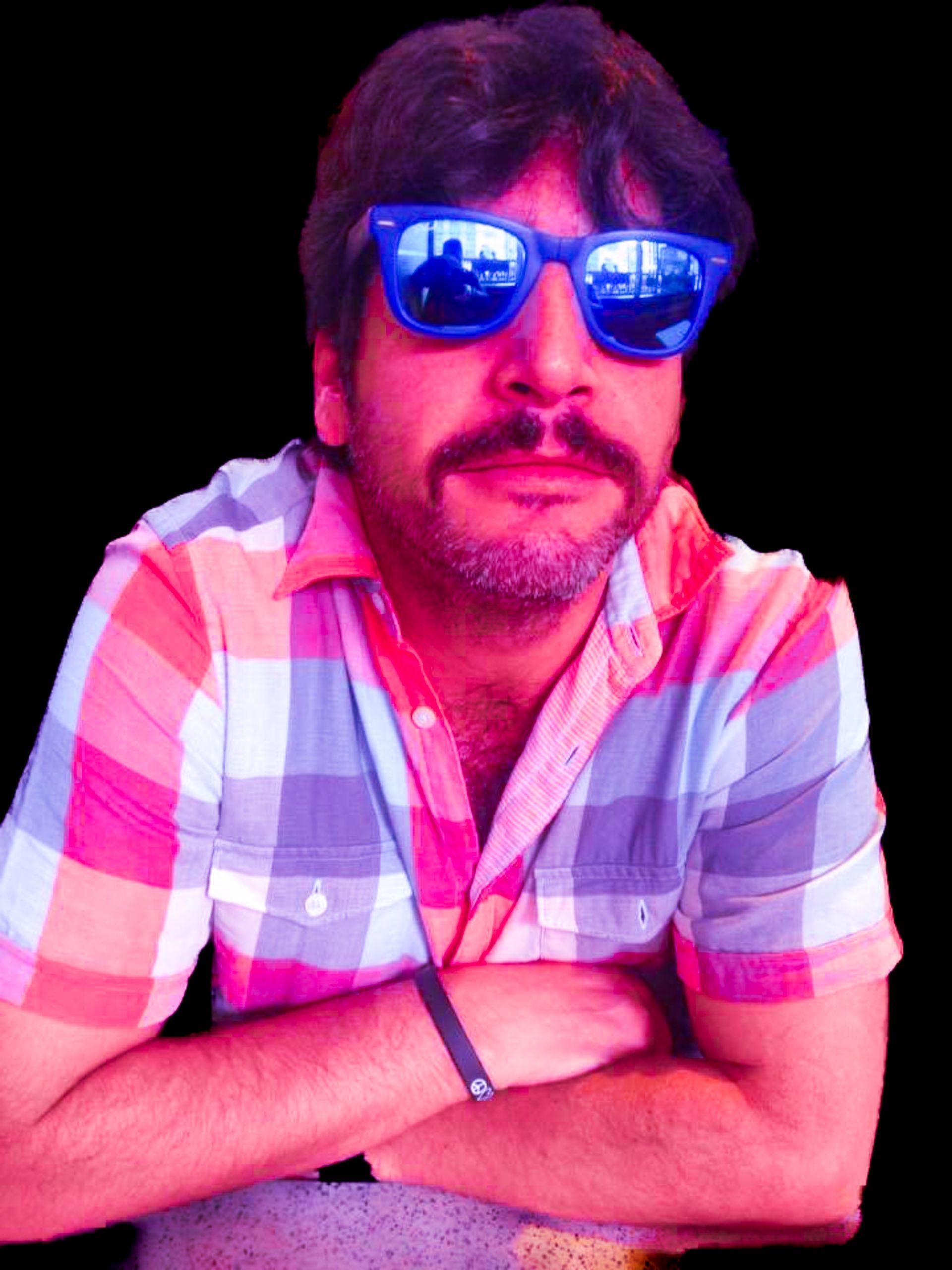 Man in blue sunglasses and a pink plaid shirt, arms crossed against a black background.