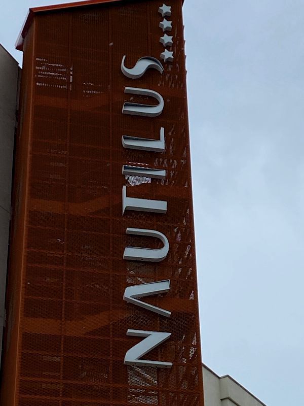 Insegna verticale NAUTILUS sulla facciata di un edificio rosso, sullo sfondo di un cielo nuvoloso.