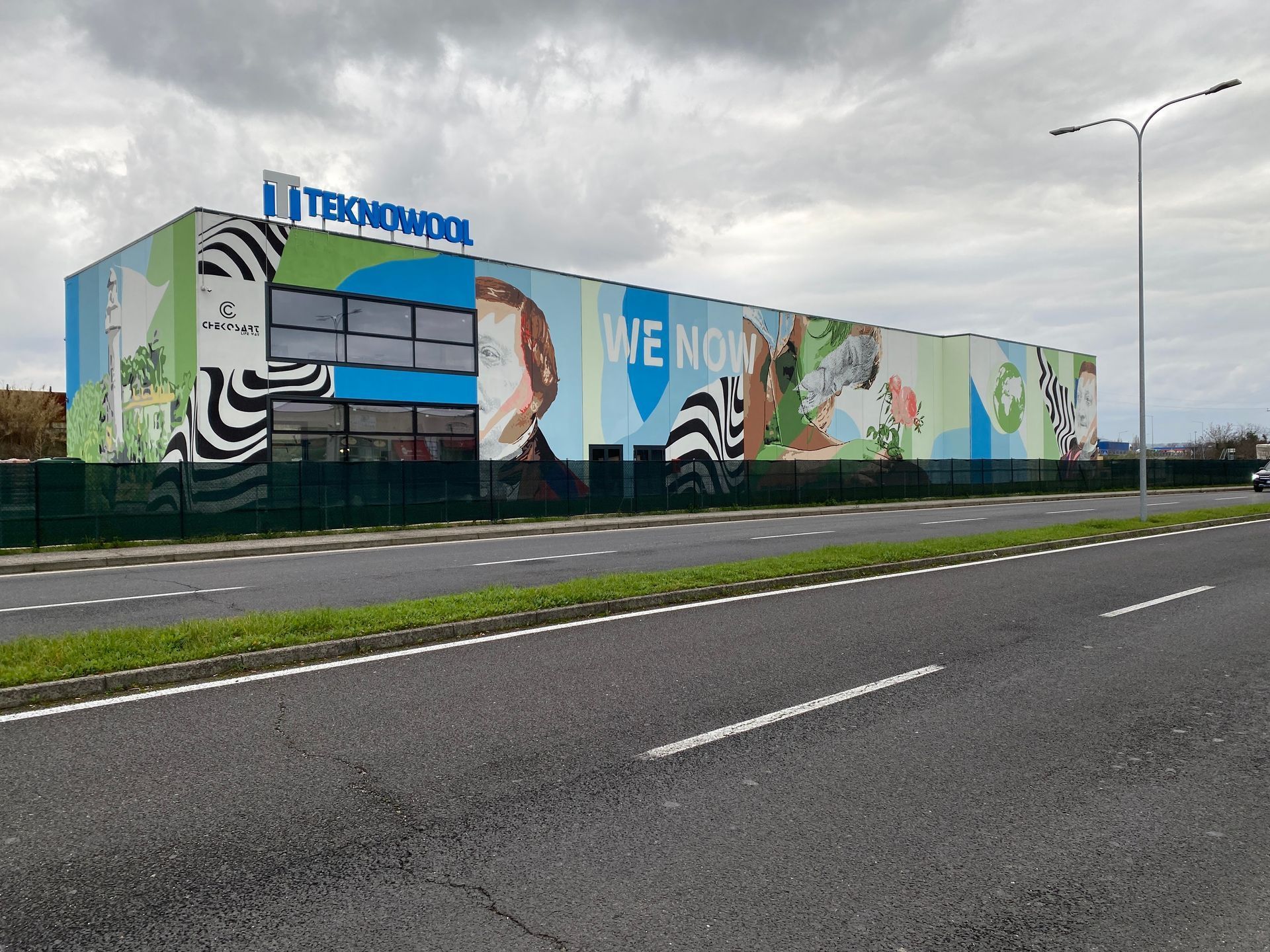 Edificio industriale con un murale colorato a lato di una strada sotto un cielo nuvoloso.