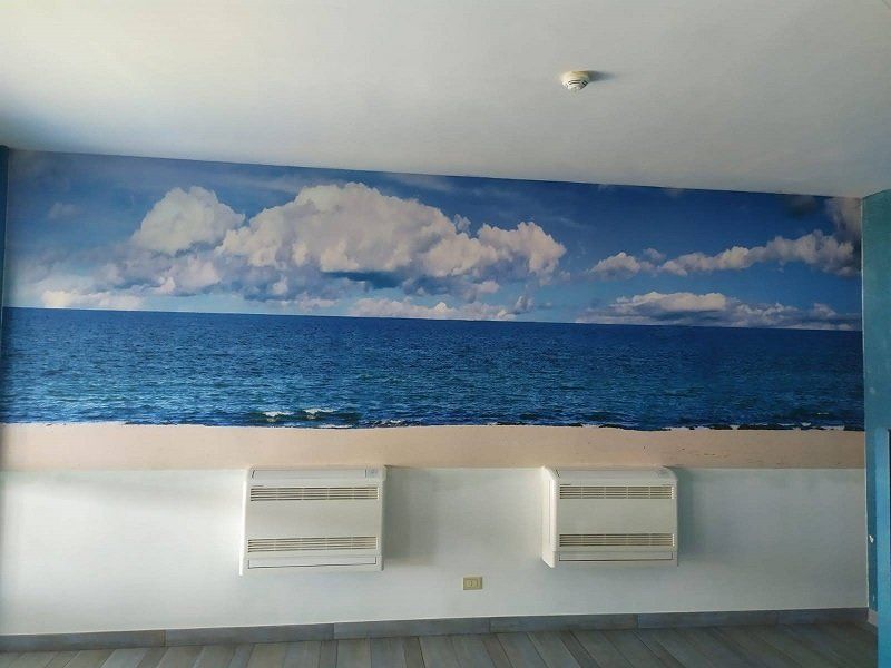 Murale raffigurante un oceano blu e un cielo nuvoloso sopra due bocchette di ventilazione a muro in una stanza.