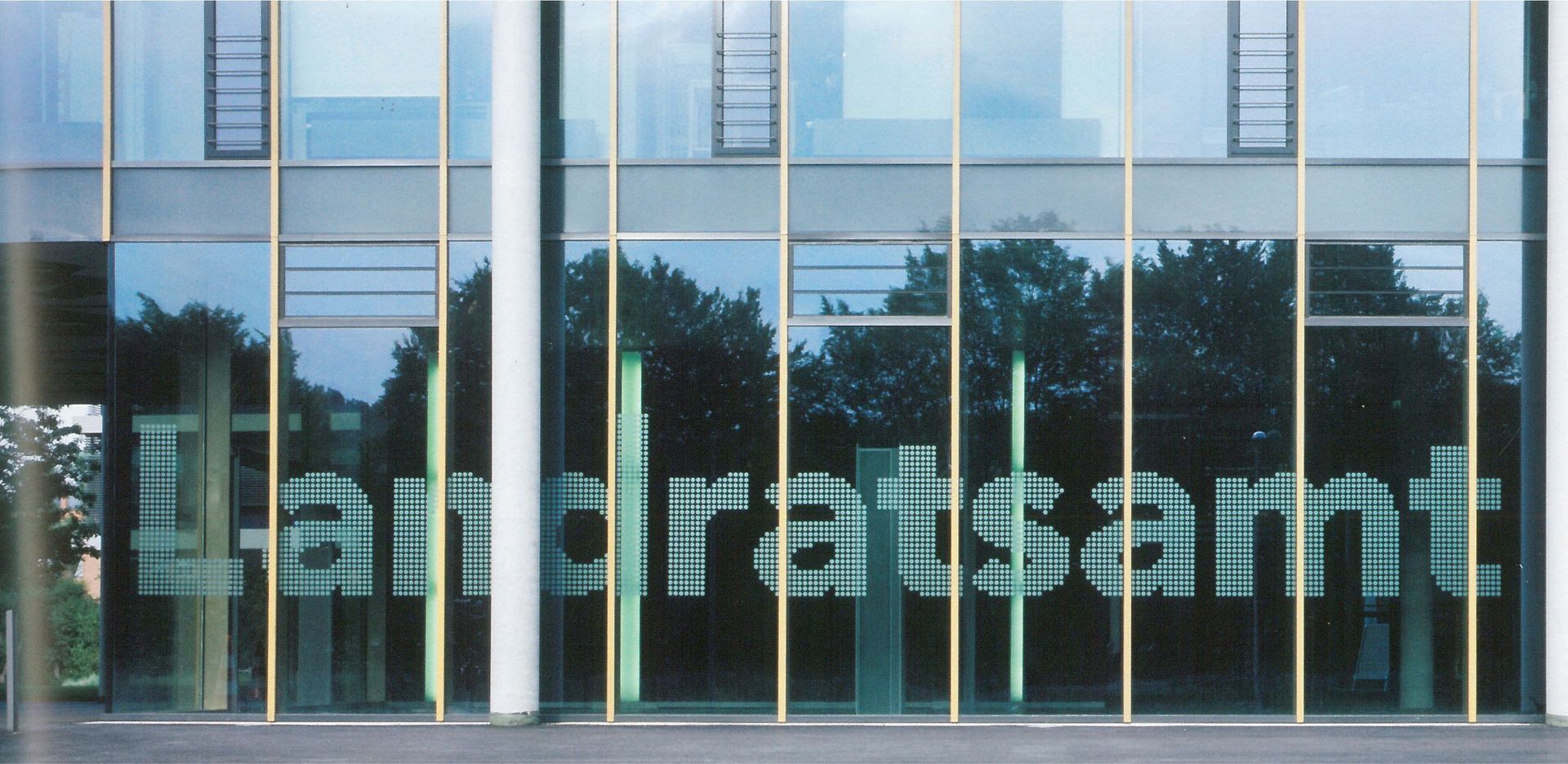 Facciata di un edificio per uffici in vetro con grandi lettere satinate e riflessi di alberi e finestre.