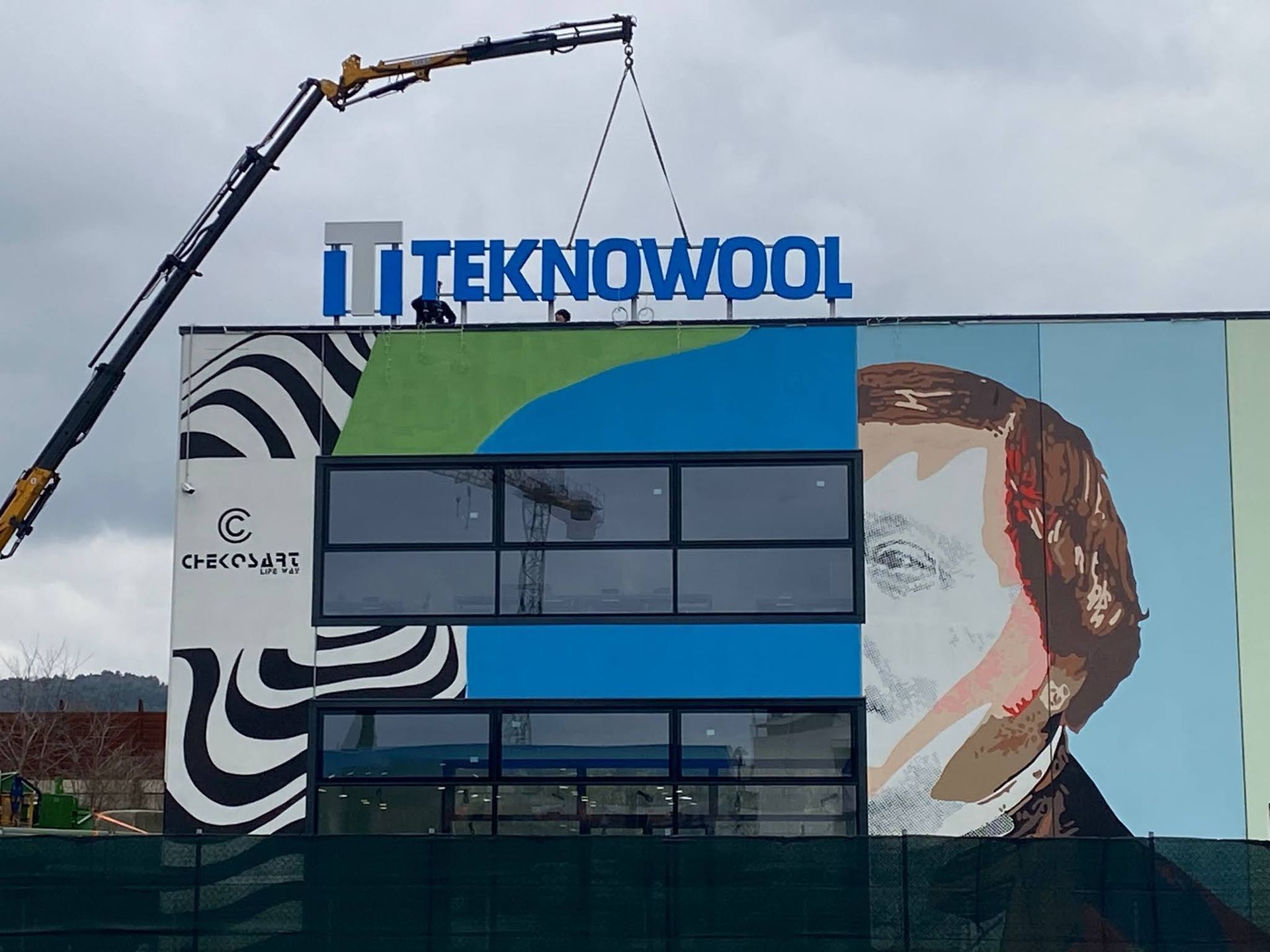 Murale dell'edificio TEKNOWOOL con insegna blu e una gru che solleva oggetti vicino al tetto