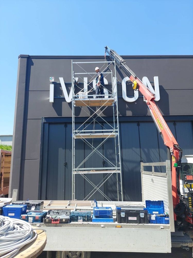 Alcuni operai, a bordo di una piattaforma elevatrice, installano un'insegna bianca sulla facciata scura di un negozio.