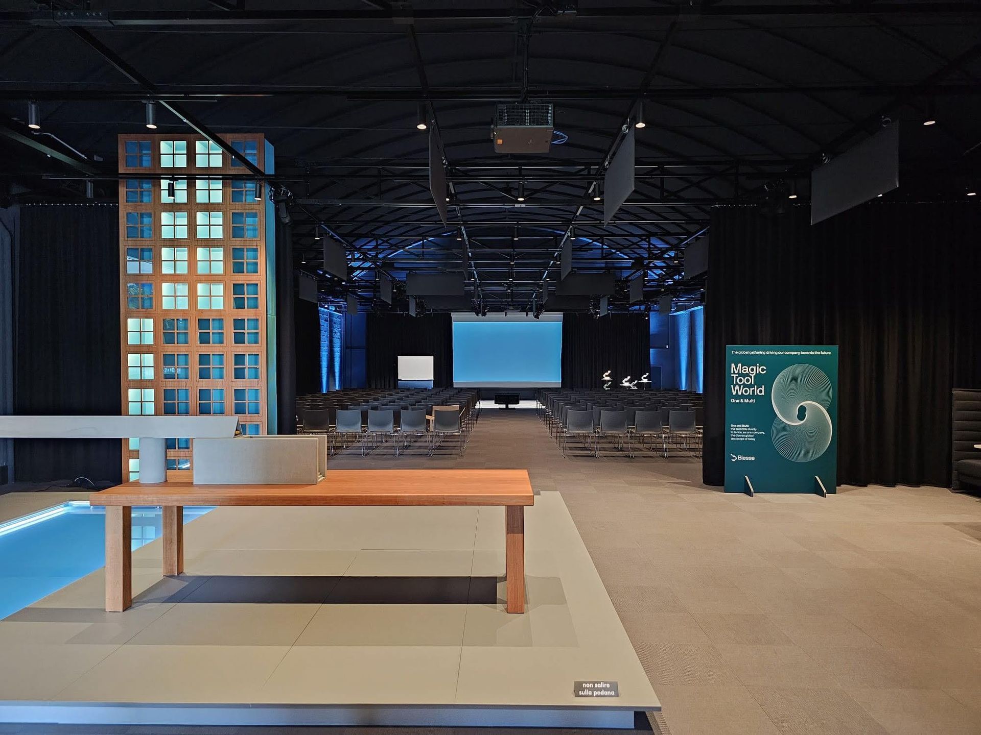 Sala eventi vuota con palco, schermo per proiezioni, file di sedie e un tavolo in primo piano.