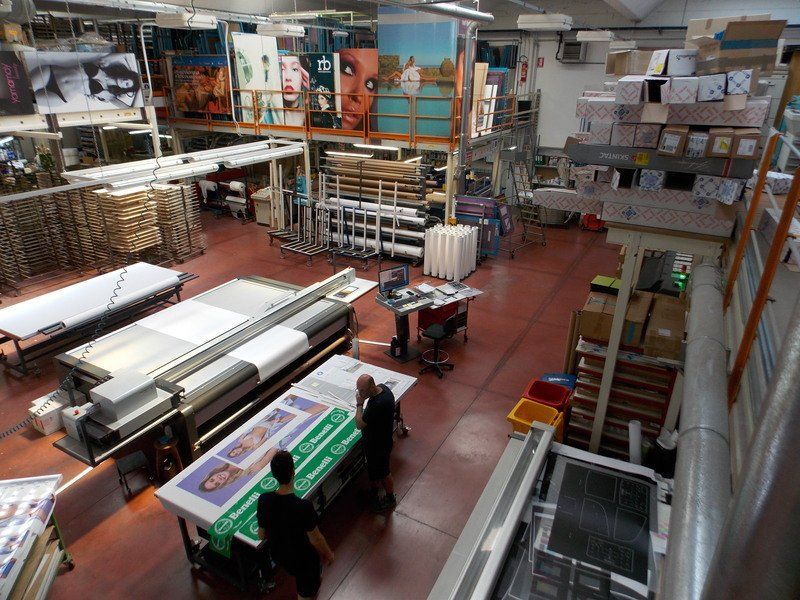 Ampia tipografia con stampanti di grande formato, materiali impilati e due operai che maneggiano uno striscione verde.