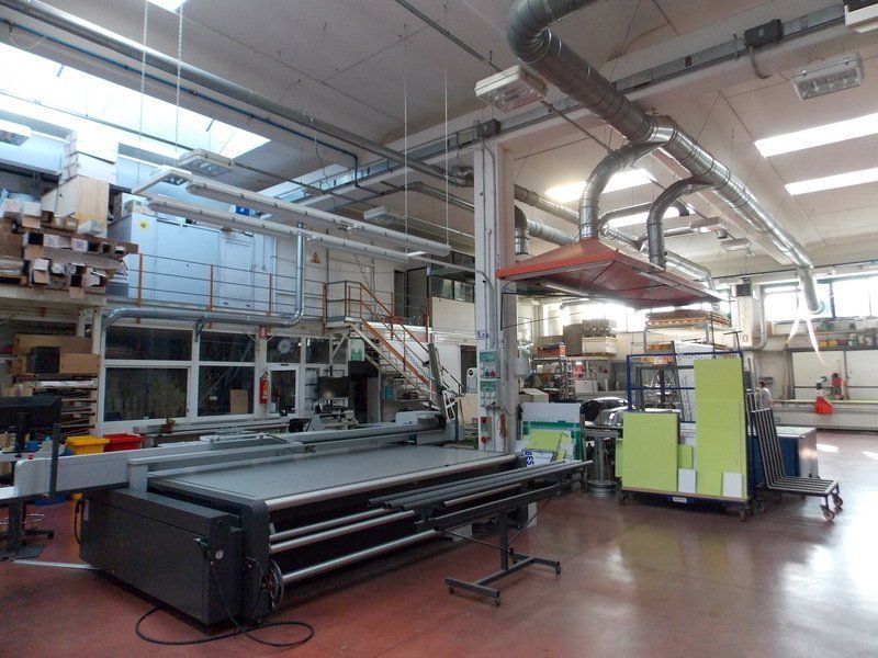 Officina industriale con grandi macchinari, condotti metallici e luci intense a soffitto in un ampio stabilimento.