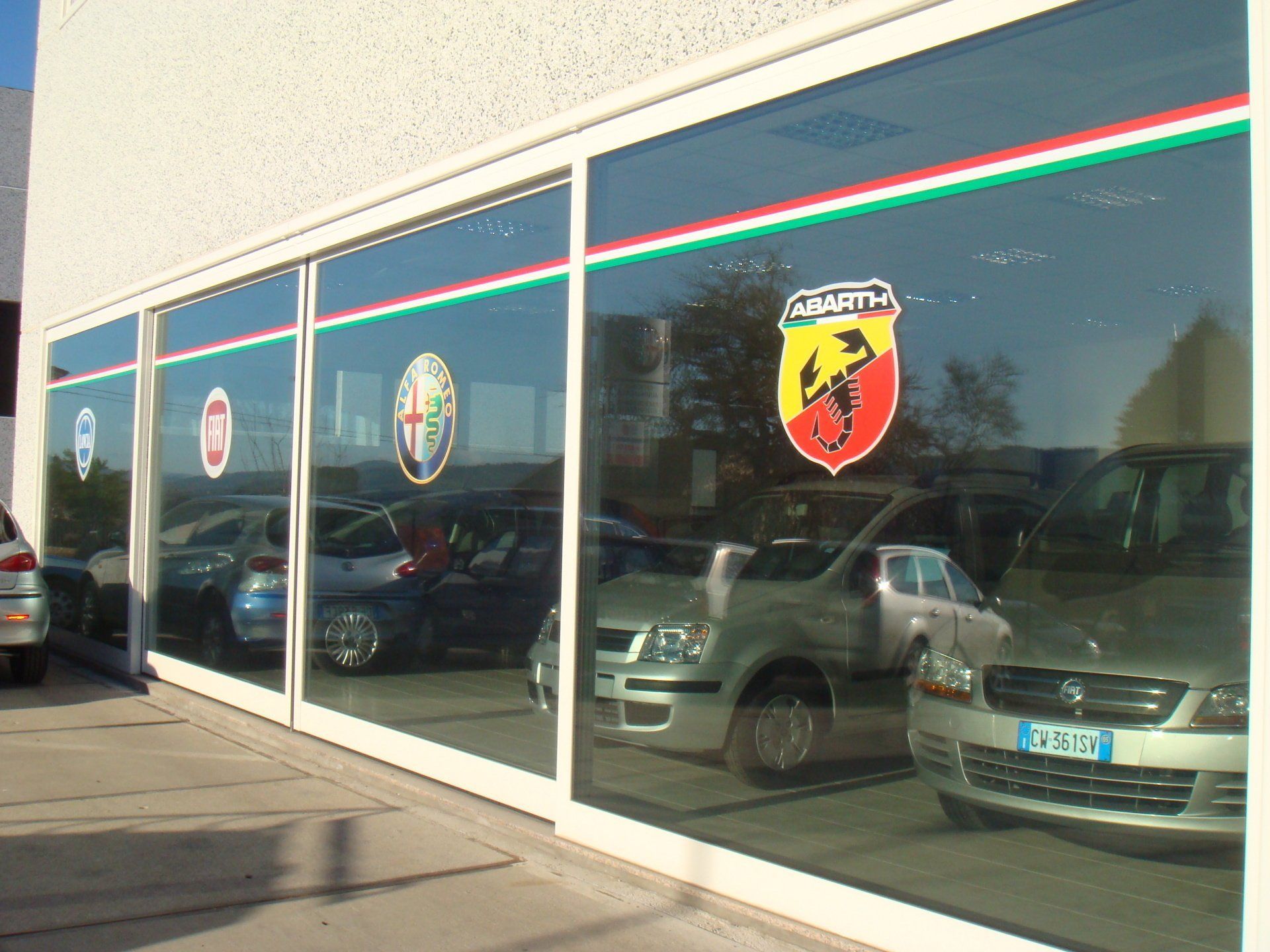 Una fila di vetrine di una concessionaria d'auto con le vetture parcheggiate che si riflettono nel vetro e un adesivo con il logo Abarth.