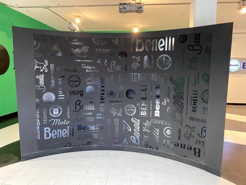 Parete espositiva curva nera con diverse grafiche di loghi bianchi in una luminosa hall interna.