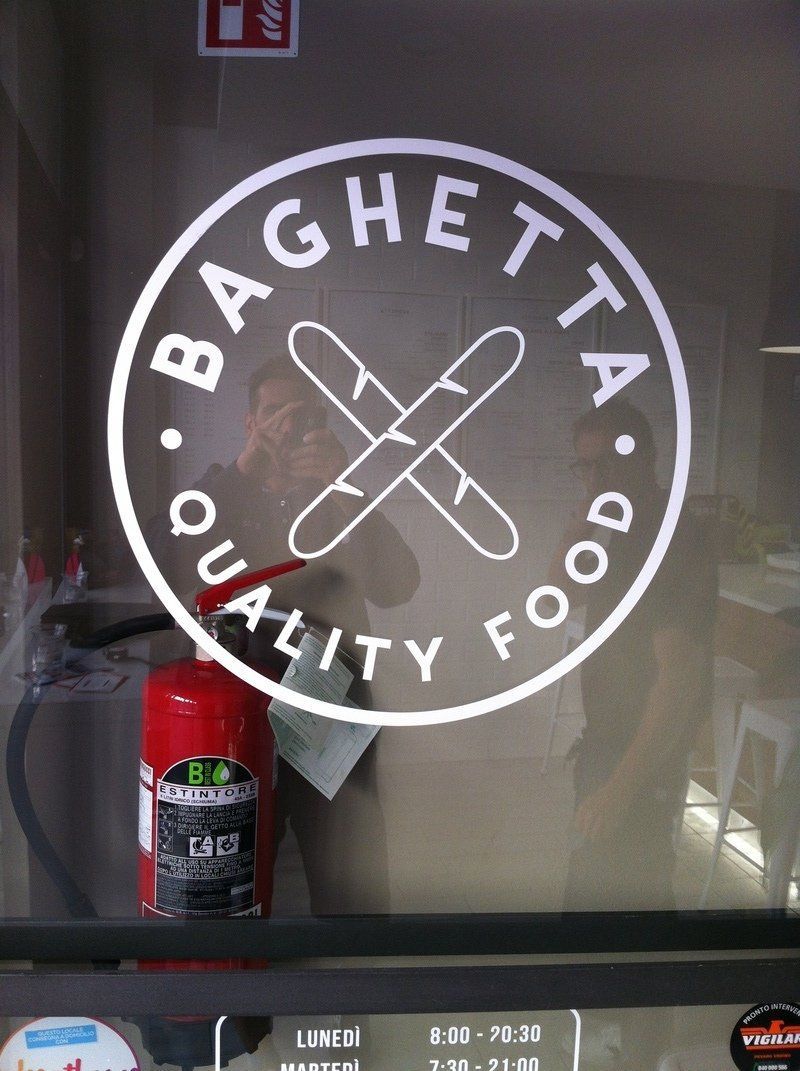 Il logo Baghetta Quality Food è posizionato su una porta a vetri, sopra un estintore rosso.