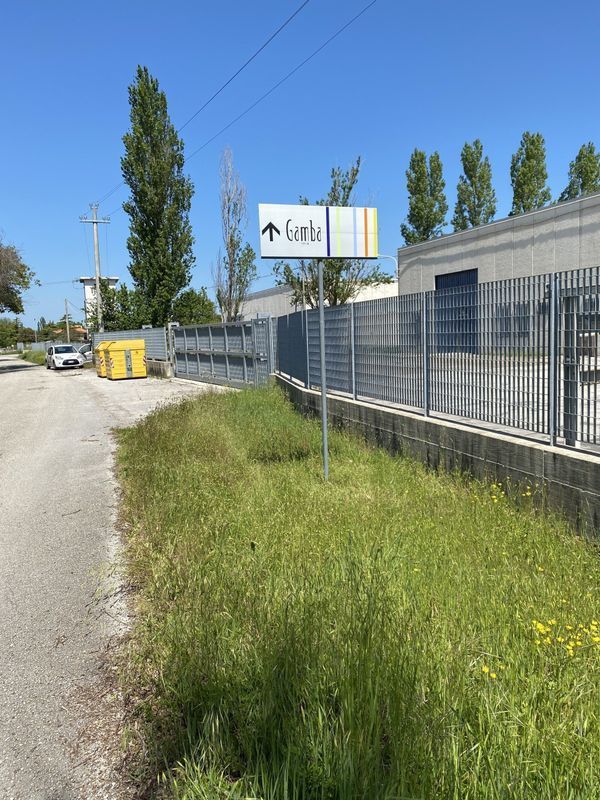 Lungo una strada rurale, accanto a un edificio industriale recintato, con un'insegna Gall e erba alta sotto un cielo azzurro e limpido.