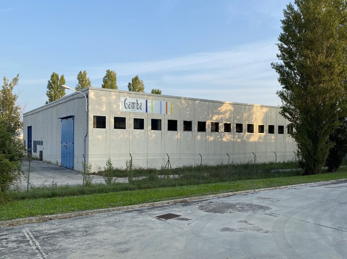Edificio industriale con porte blu e piccole finestre, adiacente a una strada asfaltata e ad alberi.