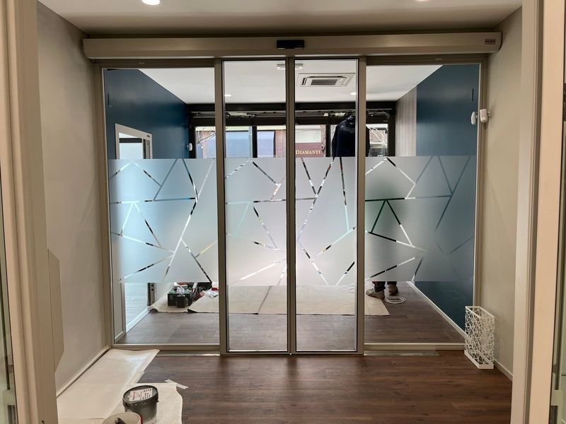 Ingresso dell'ufficio in vetro con pannelli geometrici satinati e pareti blu, visto da un corridoio.