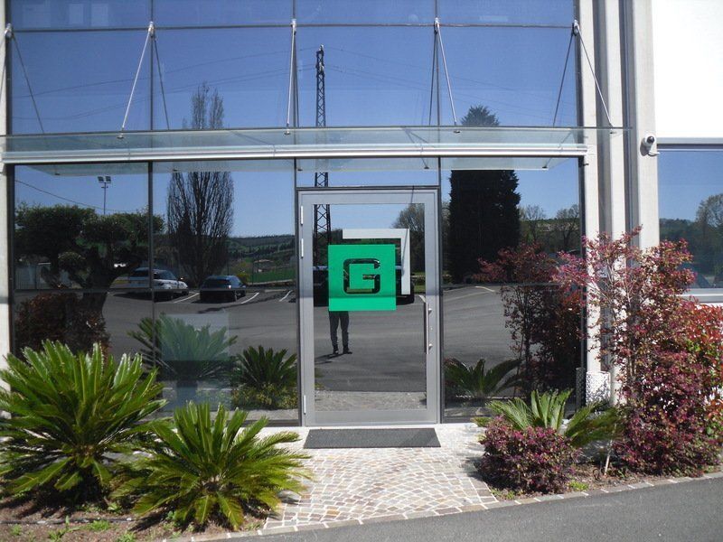 Ingresso dell'ufficio in vetro con logo verde, che riflette gli alberi e le auto all'esterno.