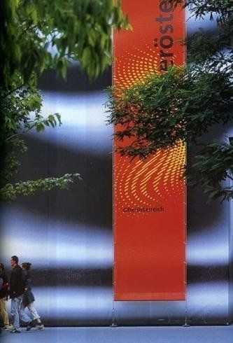 Striscione verticale arancione su uno sfondo sfocato, con alberi e due persone in basso a sinistra.