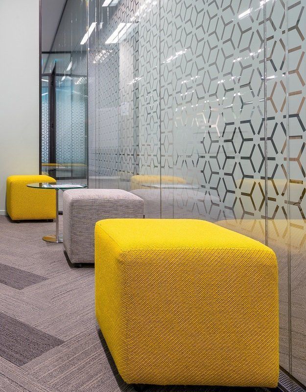 Moderna area relax per ufficio con sedute cubiche gialle e grigie accanto a una parete divisoria in vetro con finitura satinata geometrica.