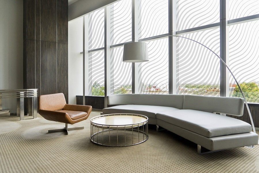 Salotto moderno con divano grigio curvo, poltrona color cuoio, tavolino rotondo e grandi finestre decorate.