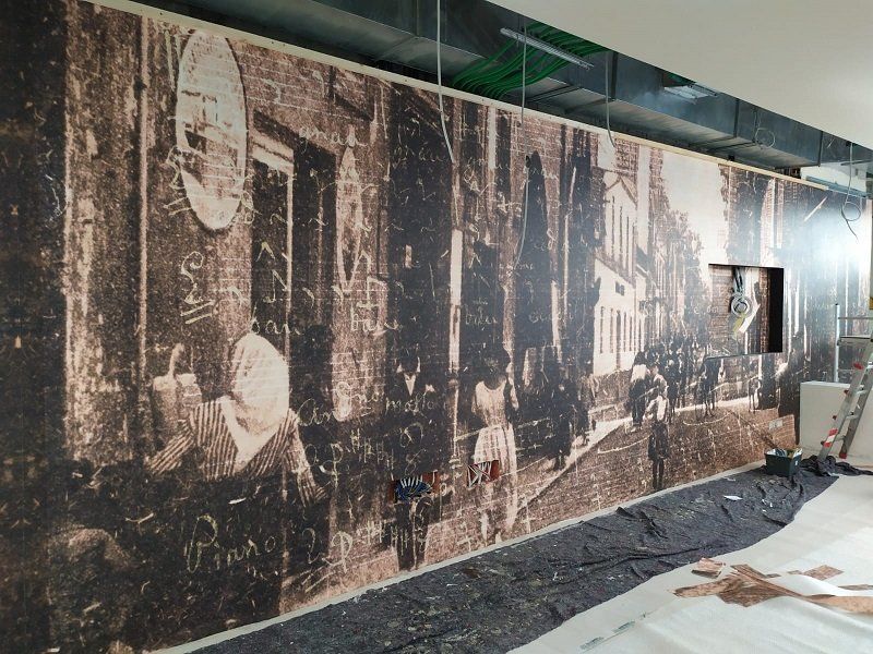 Grande murale color seppia su una parete raffigurante una vecchia strada cittadina con edifici e pedoni.