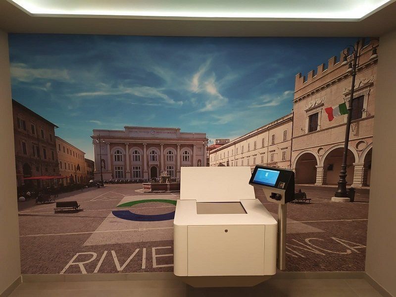 Esposizione museale con un chiosco bianco di fronte a un grande murale raffigurante una piazza di una città italiana.