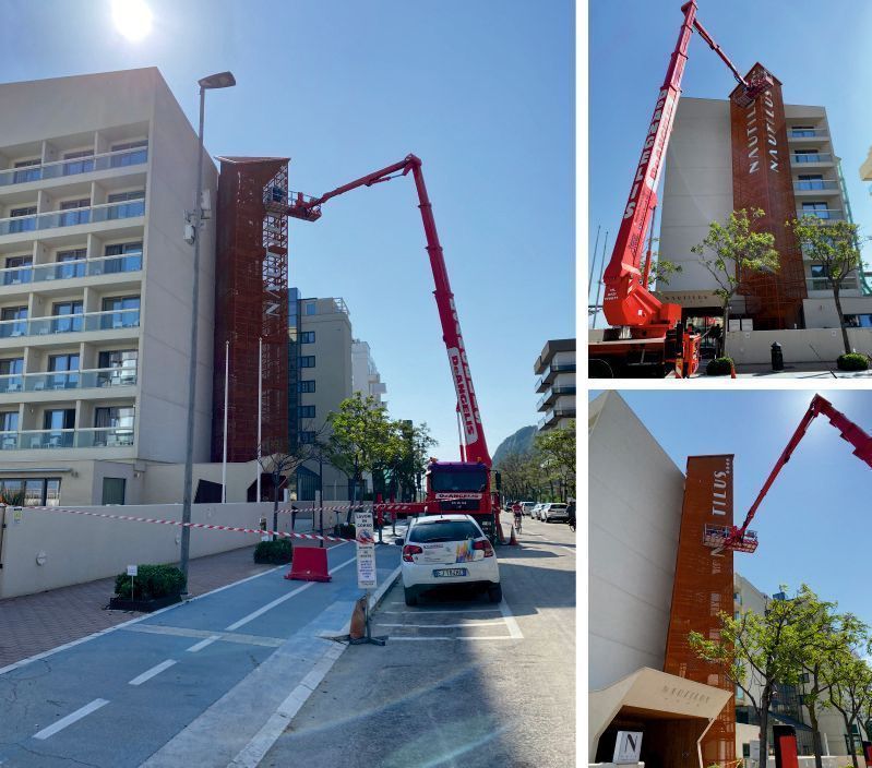 Un camion con gru rosso è parcheggiato accanto a un condominio e solleva dei materiali in una strada soleggiata con auto parcheggiate.