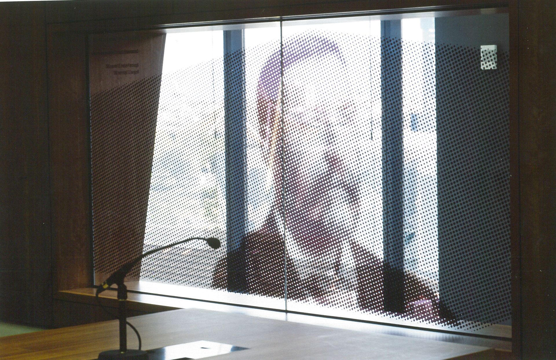 Una persona parla dietro uno schermo a rete in un'aula di tribunale, con un microfono in primo piano.