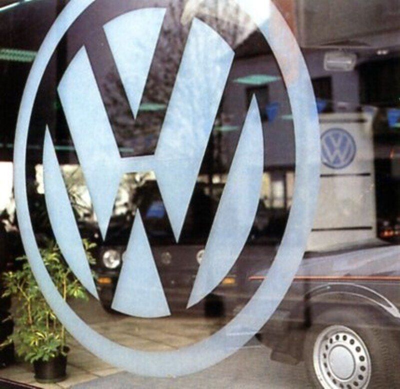 Logo Volkswagen su una vetrina, con un veicolo VW visibile sullo sfondo.
