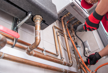 A technician in red work clothes wearing gloves uses a tool to repair copper plumbing pipes near a wall-mounted boiler.