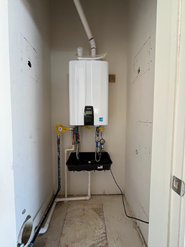 A Navien tankless water heater mounted on a gray wall with copper plumbing pipes, isolation valves, and white vent pipes.
