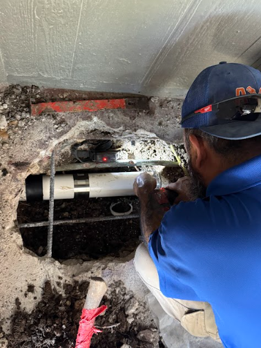A plumber wearing grey gloves uses an orange pipe wrench to tighten a metal drain pipe under a sink.