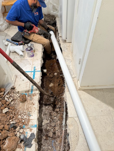 A copper plumbing pipe has multiple pinhole leaks, spraying water onto a wooden floor joist in a crawl space.