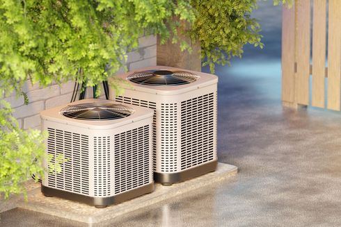 Two outdoor HVAC units beneath a tree next to a walkway