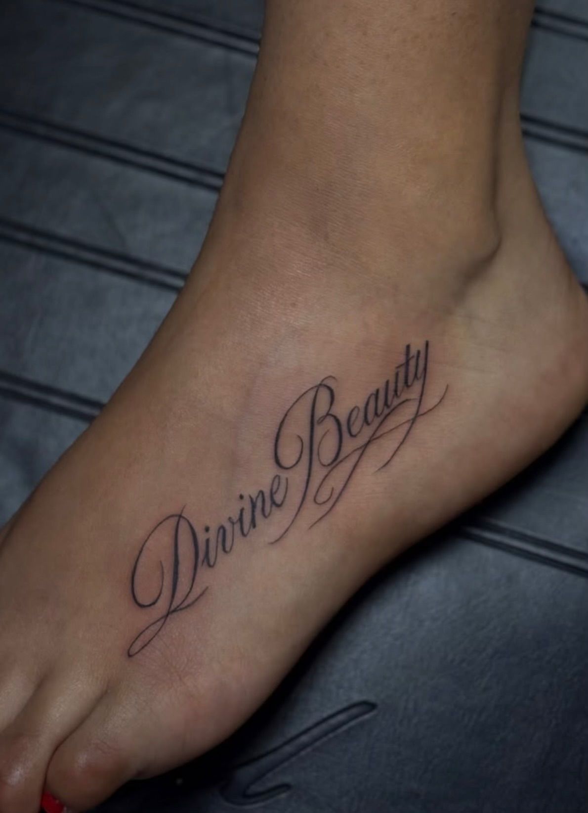 A fine-line script tattoo reading Divine Beauty across the top of a foot