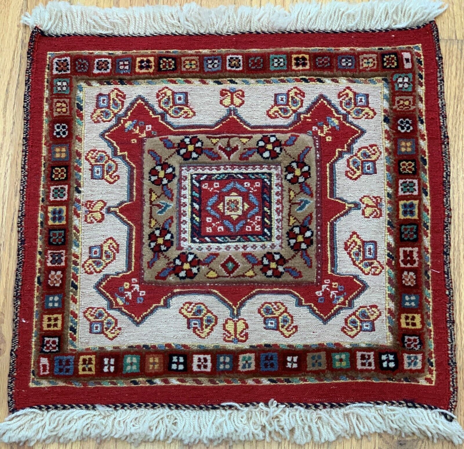Red, cream, and blue patterned square persian rug with fringe on a light background