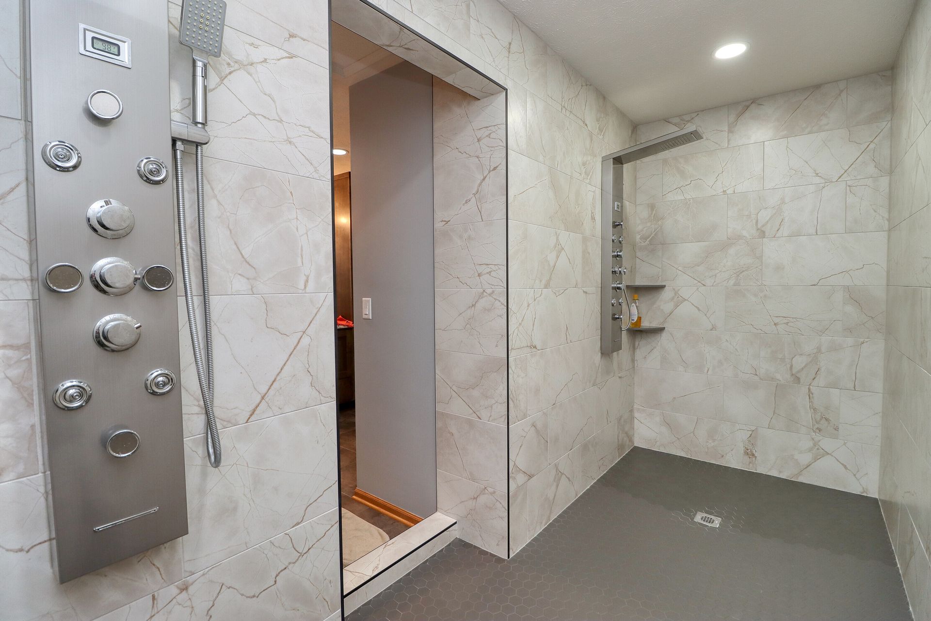 Modern marble-tiled shower with stainless-steel control panel and open doorway to a lit bathroom.
