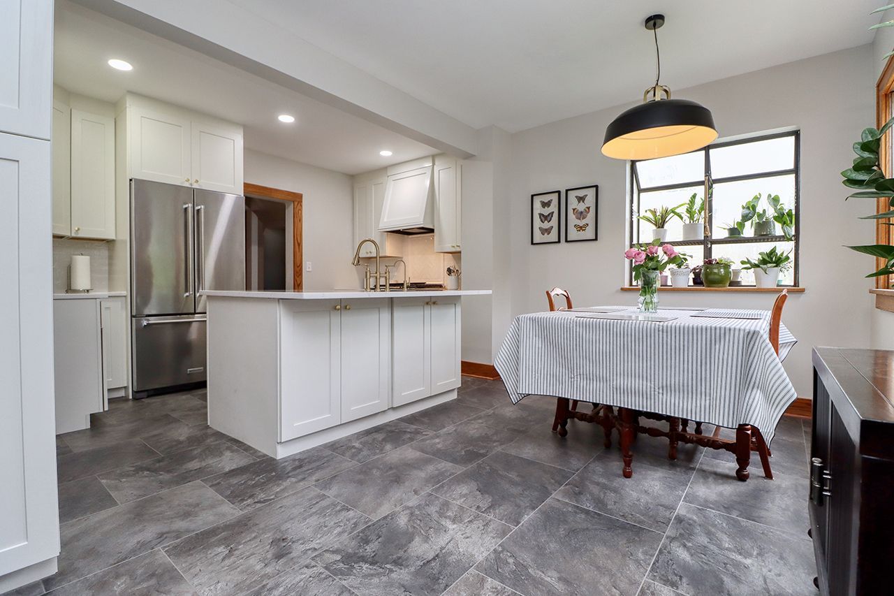 Modern kitchen and dining area with white island, gray tile floor, and a table by a window.